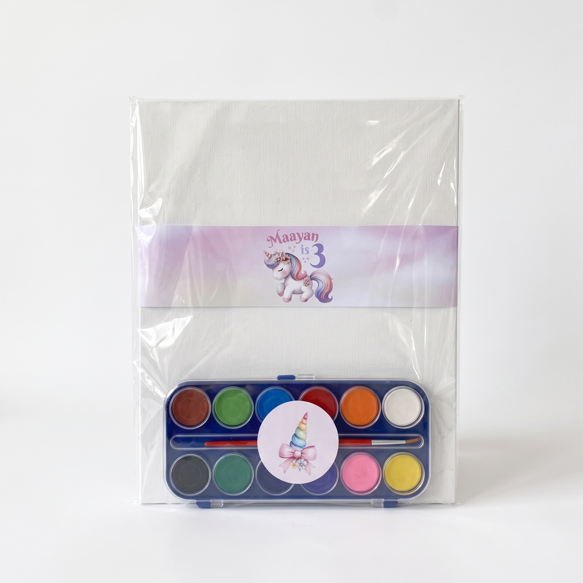 A children's watercolor painting set in a clear plastic sleeve, featuring a blue palette with 12 colorful paint wells, a paintbrush, and a round sticker with a unicorn illustration. A purple personalized band reading 'Maayan 3' with a unicorn is wrapped around the package.