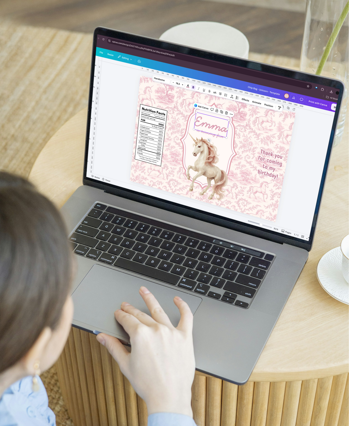 A woman in a light blue shirt uses a silver laptop on a round wooden table, looking at a pink digital design template featuring a unicorn.