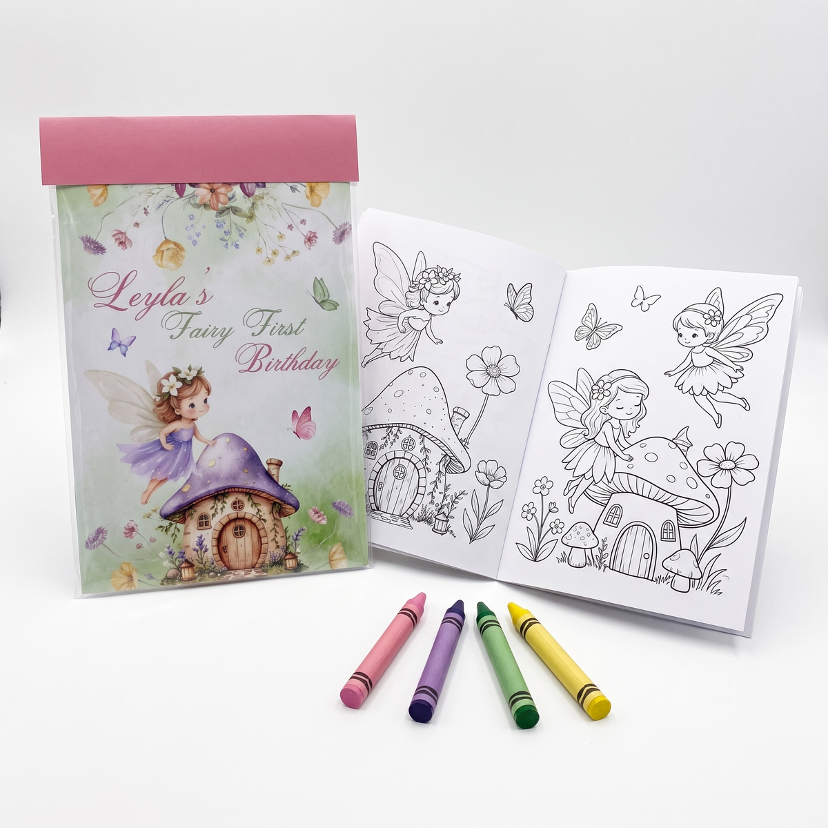 A personalized fairy-themed coloring book set shown open and closed, with four crayons in pink, purple, green, and yellow placed in front on a clean white background.