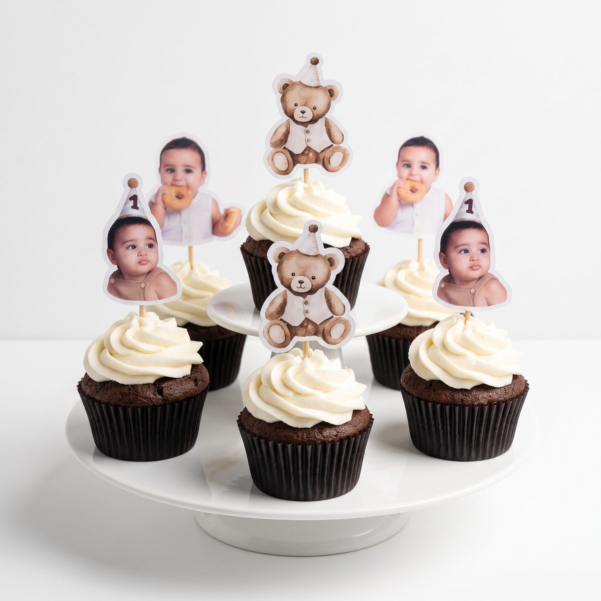 A set of custom photo cupcake toppers displayed on a white cupcake stand. The toppers feature cute designs including a teddy bear wearing a party hat and small photo cutouts of babies with '1' birthday flags, decorated on top of chocolate cupcakes with white frosting.