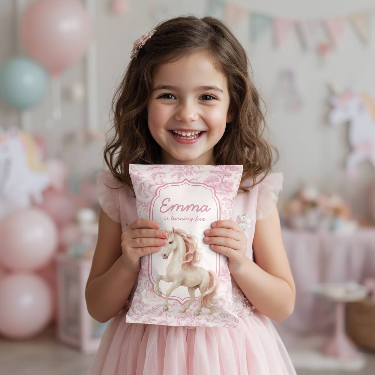 A young girl with brown hair smiles while holding a pink personalized party snack bag featuring a unicorn illustration and the name 'Emma'. The background shows a softly blurred pink and white unicorn-themed party setup with balloons and bunting.
