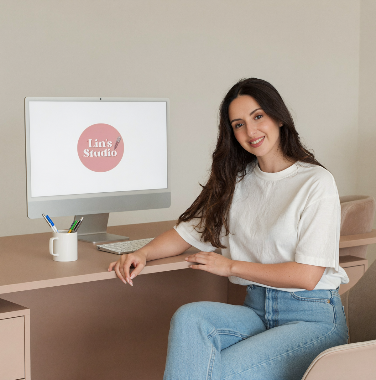A woman with long dark hair sits in a beige chair at a light wood desk, smiling at a computer monitor displaying the logo 'Lins Studio'. She is wearing a white t-shirt and blue jeans in a bright, modern home office setting.