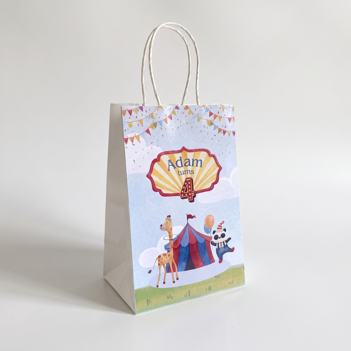 A white paper gift bag with twisted paper handles, featuring a colorful circus-themed illustration with the text 'Adam 4' and cartoon animals on a light gray background.