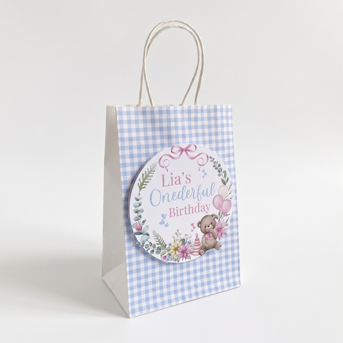 A small paper gift bag with a light blue and white gingham pattern, featuring a custom round sticker that reads 'Lia's One Dexter Birthday' with a teddy bear and floral design, set against a plain white background.