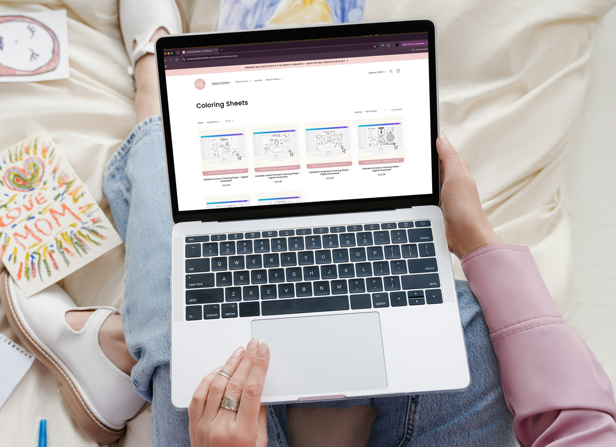 A person sitting on a bed holds a silver laptop showing a website page, surrounded by colorful children's drawings, a marker, and a card on a cream-colored bedspread.